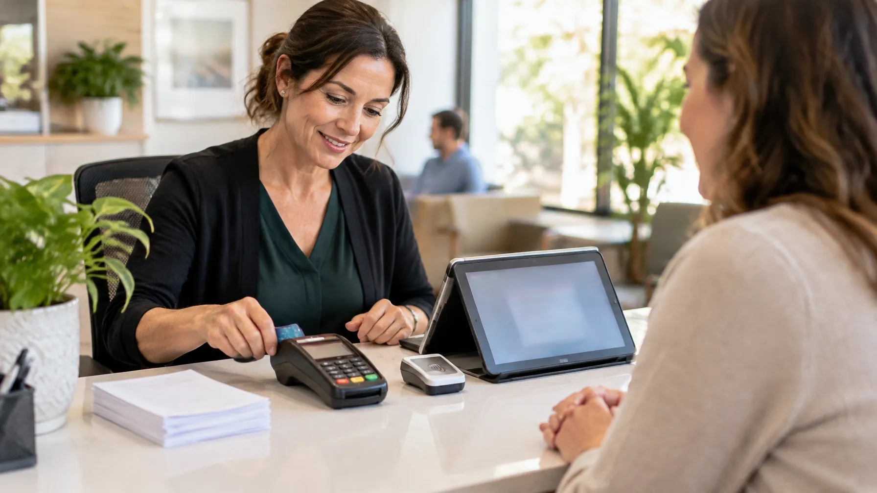 A practice manager processing a card payment at a front desk with a payment terminal and tablet.
